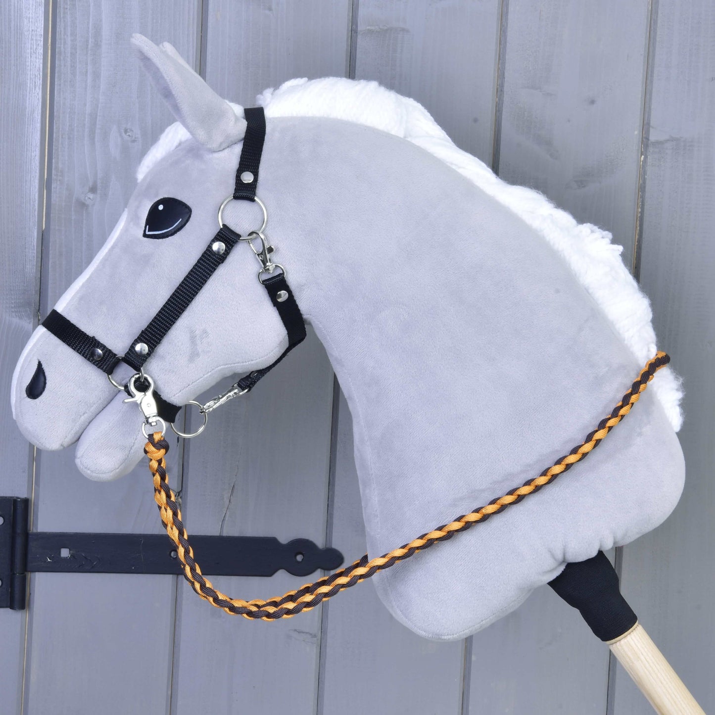 Pletené opraty Knitted Pomello od Hobby Horse LarDen na sivom Hobby Horse s bielou hrivou a čiernou uzdečkou — pletené polypropylénové opraty v oranžovej a hnedej farbe — nasadenom na drevenej tyči pred sivým dreveným pozadím.
