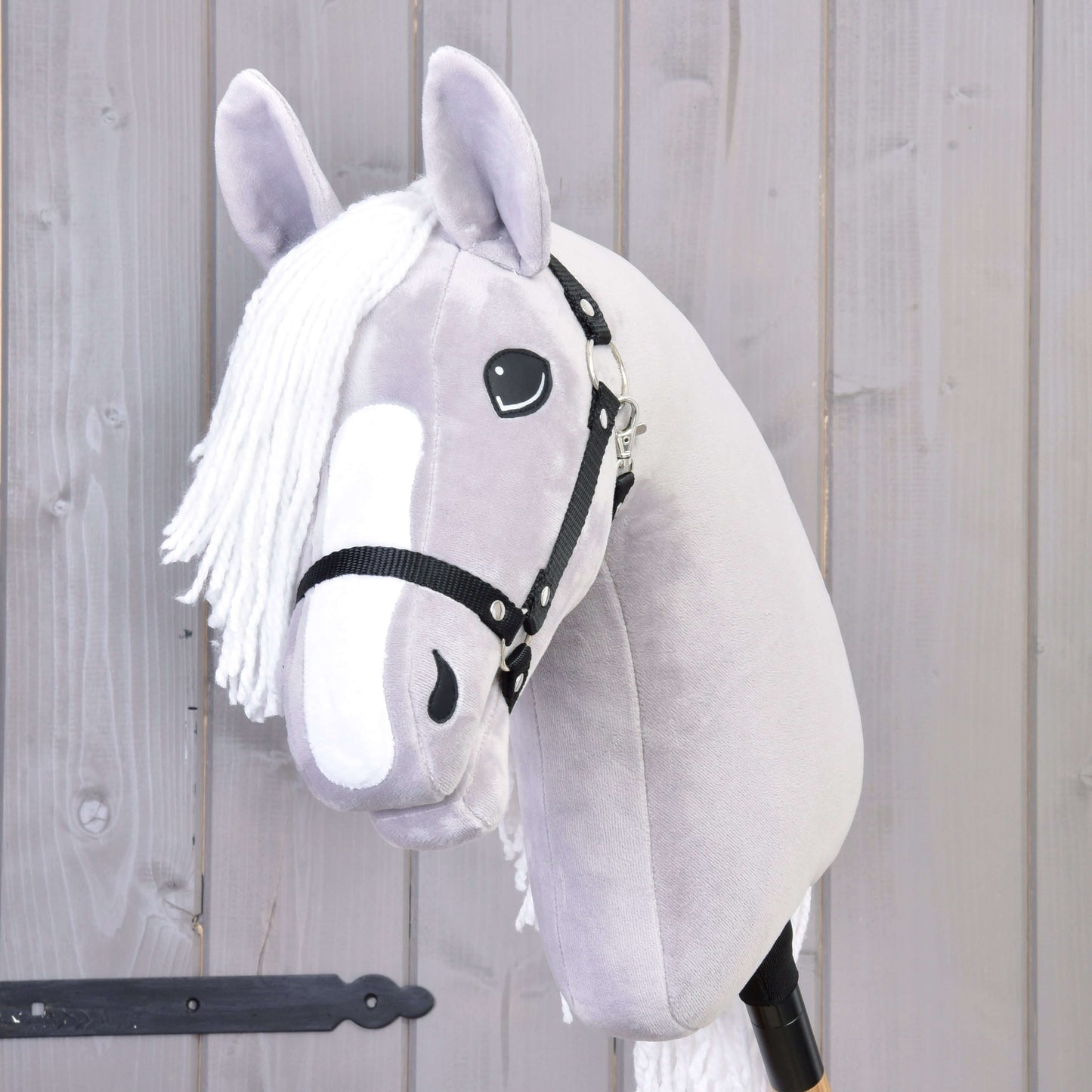 Hobby Horse Popcorn s čiernou ohlávkou od Hobby Horse LarDen, so sivým plyšovým prevedením, bielou hrivou a priateľskou tvárou – perfektný spoločník pre nápaditú hru. Zobrazený pred svetlou drevenou stenou.