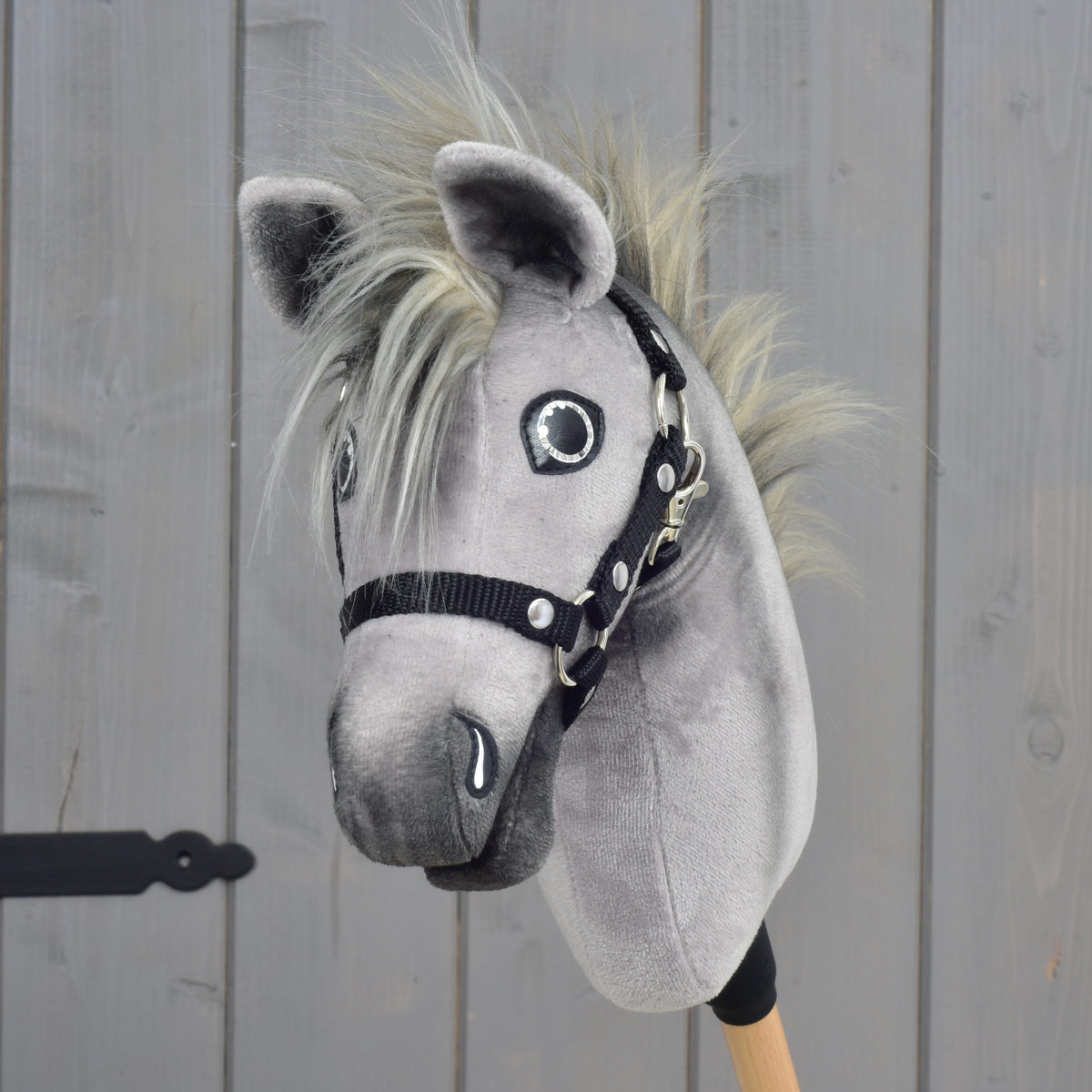 Hobby Horse Prestige s čiernou ohlávkou, veľkosť XS od Hobby Horse LarDen so sivým plyšovým telom, blond hrivou a drevenou tyčou, zobrazený pred svetlohnedou drevenou stenou.