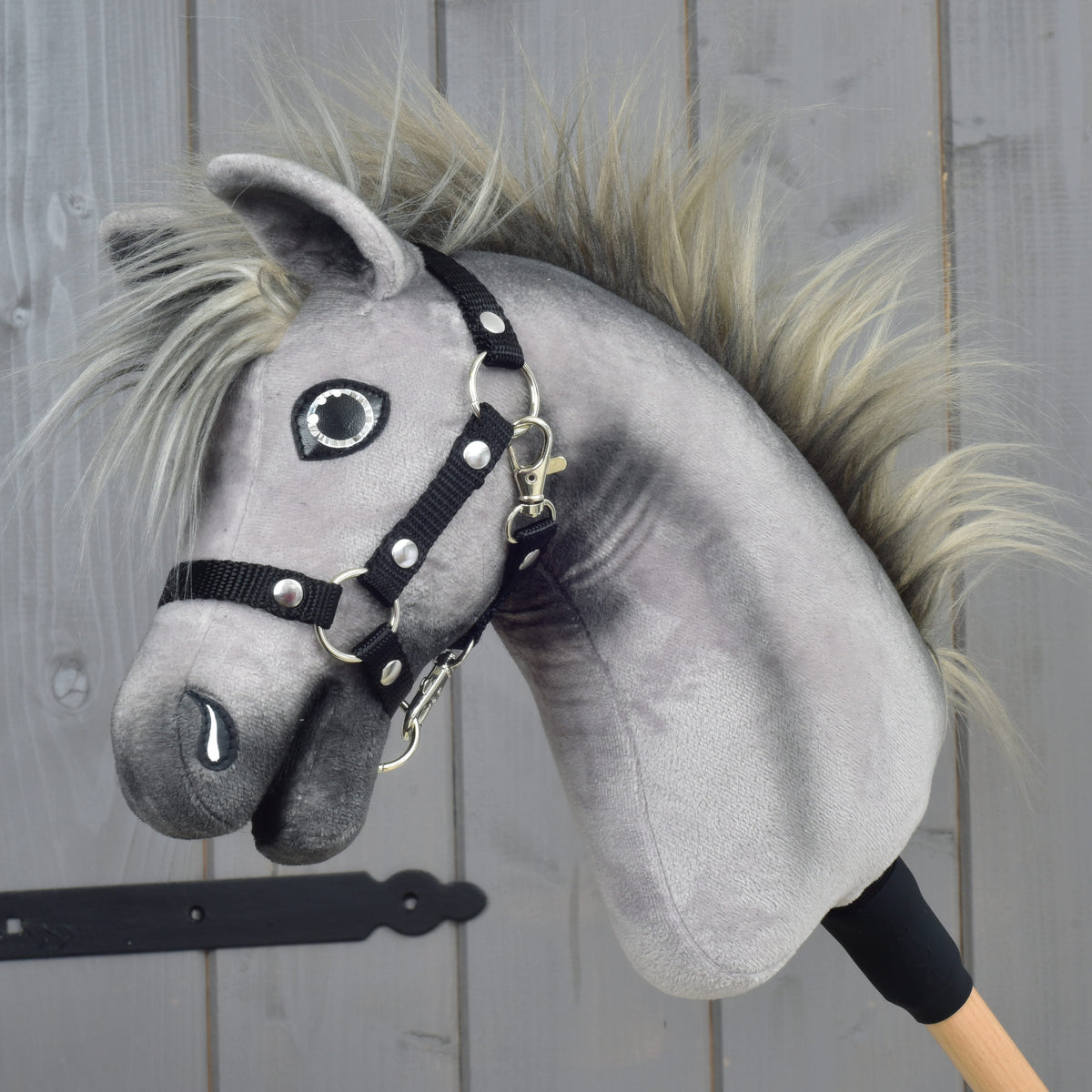 Hobby Horse Prestige s čiernou ohlávkou, veľkosť XS od Hobby Horse LarDen so sivým plyšovým prevedením, rozstrapatenou hrivou a čiernou ohlávkou, inšpirovaný eleganciou anglického teplokrvníka. Je nasadený na drevenej tyči a zobrazený na pozadí drevených panelov.