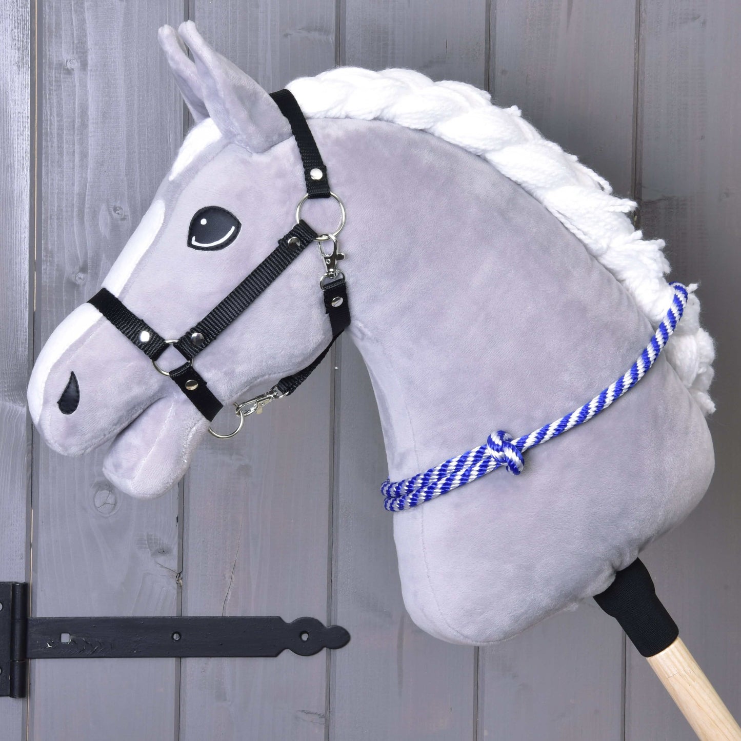 Nákrčník prúžok modrý od Hobby Horse LarDen na sivom plyšovom Hobby Horse s bielou hrivou, čiernou ohlávkou a modro pruhovaným cordeom, na drevenej tyči pred sivou drevenou stenou.