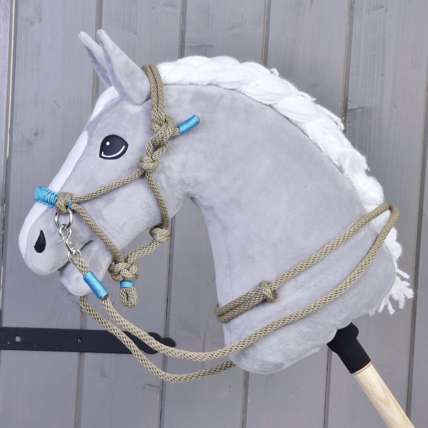 Plyšová sivá hlava Hobby Horse LarDen s bielou hrivou a ohlávkou Povrazový set Nature Blue je nasadená na drevenej tyči a umiestnená pred pozadím zo sivých drevených panelov.