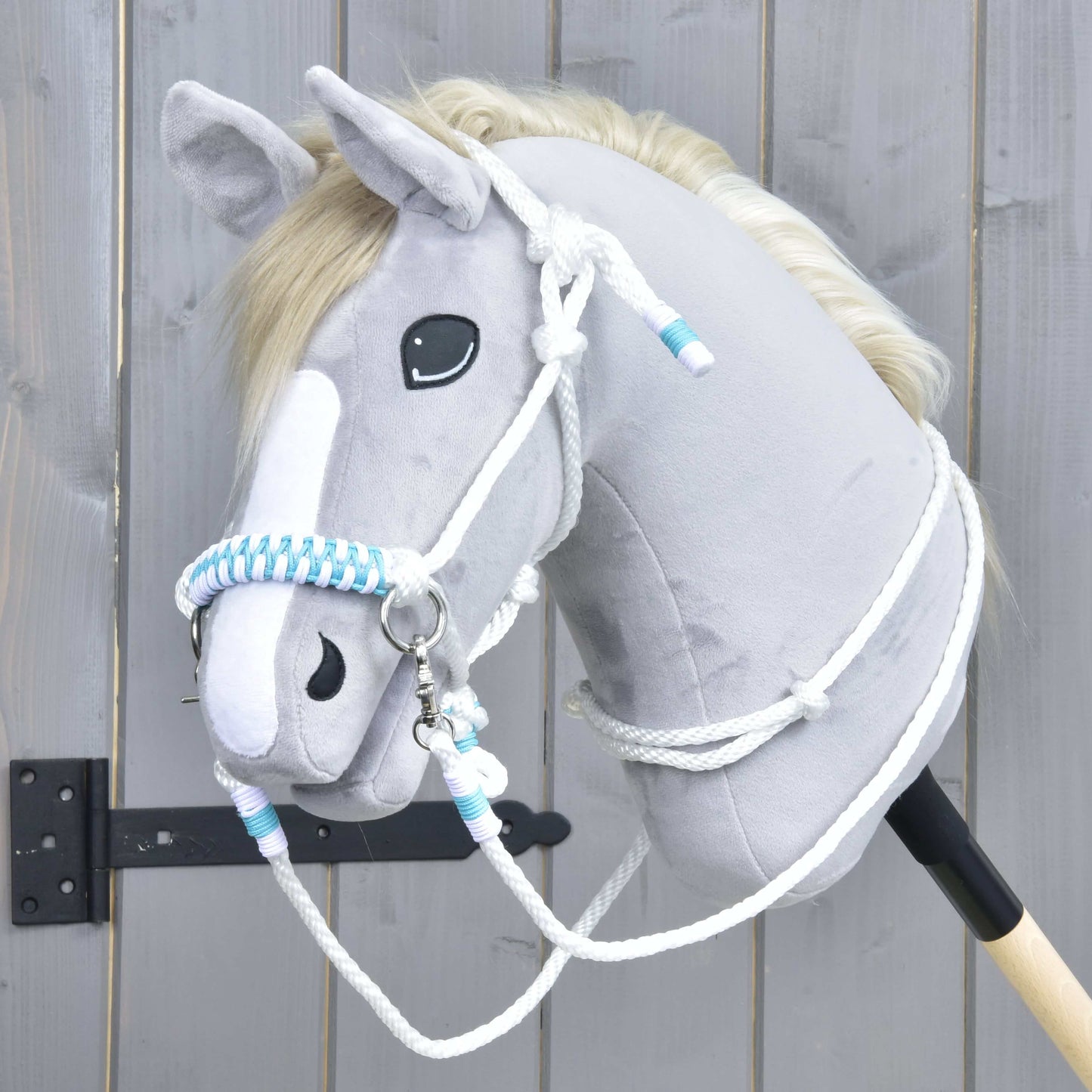 Hobby Horse LarDen Povrazový set White Queen s plyšovou hlavou Hobby Horse vo svetlosivej farbe, bielou hrivou a pruhom na tvári a modrobielo pletenými opratami, na pozadí dreveného plota.
