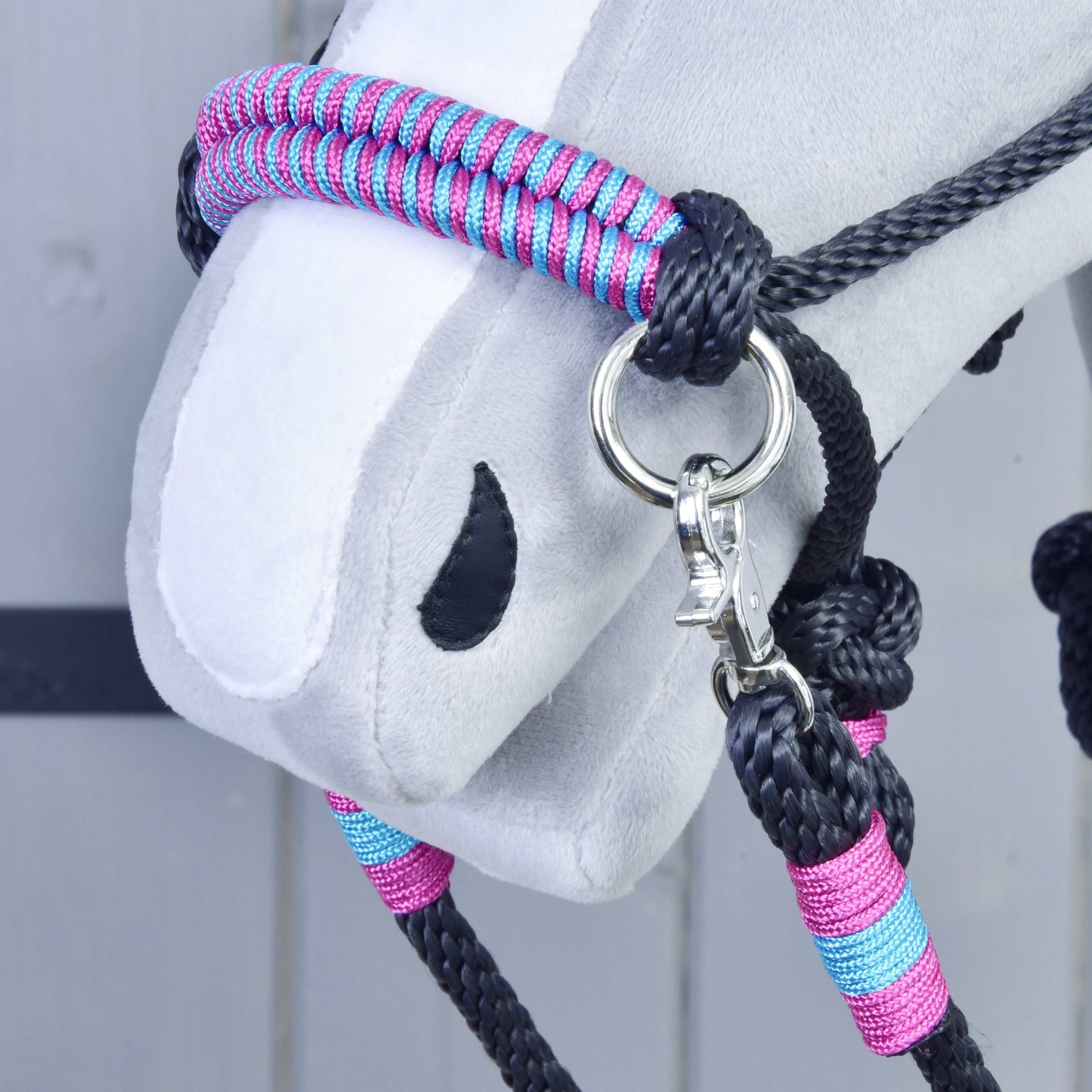 Detailný záber na Povrazový set Princess od Hobby Horse LarDen na hlave plyšového Hobby Horse – čierna, ružová a modrá pletená ohlávka s kovovými krúžkami a karabínou – ideálny pre nápadité jazdecké zostavy na svetlo sivom drevenom pozadí.