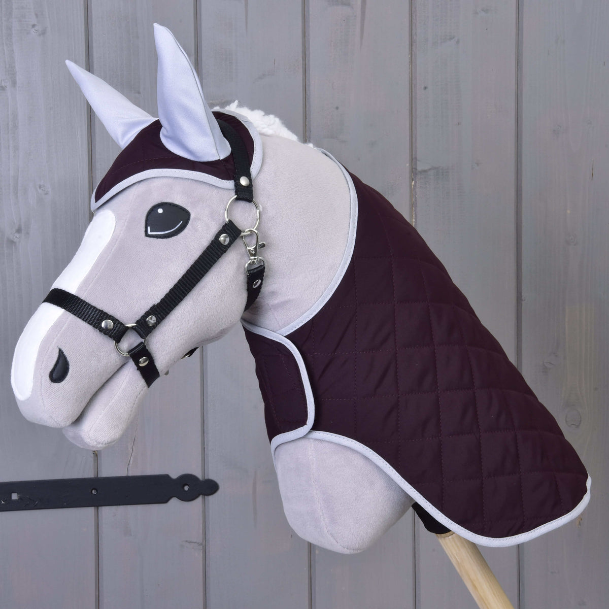 Hobby Horse LarDen Set Purple obsahuje sivú hračku Hobby Horse s prešívanou tmavofialovou dekou, uzdečkou a náušníkom, všetko nasadené na drevenej tyči a zobrazené na pozadí z drevených dosiek.
