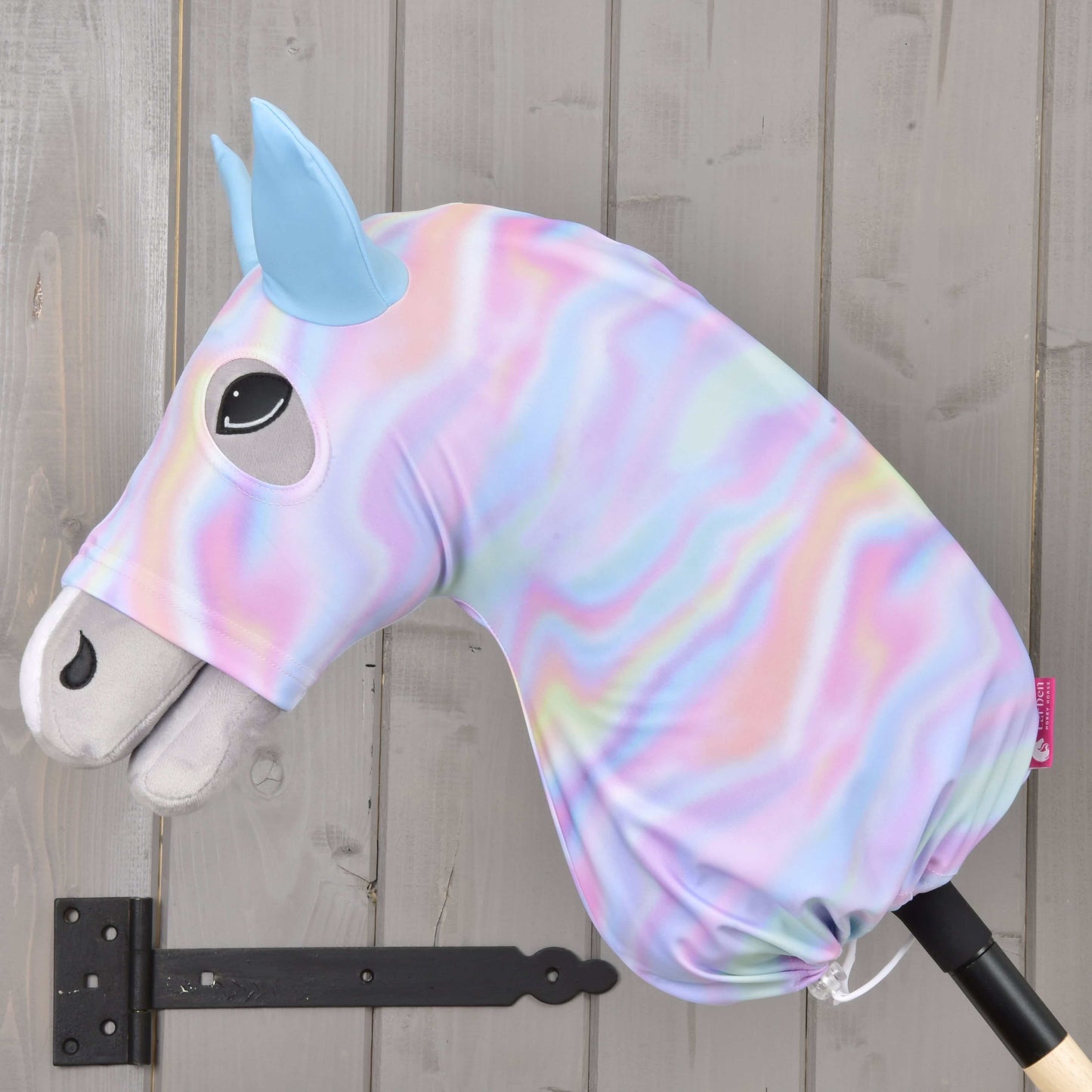 Hobby Horse ochranný oblek Rainbow od Hobby Horse LarDen z pastelové duhově spirálové lycry se světle modrýma ušima, vystavený na černém kovovém držáku před šedou dřevěnou stěnou.