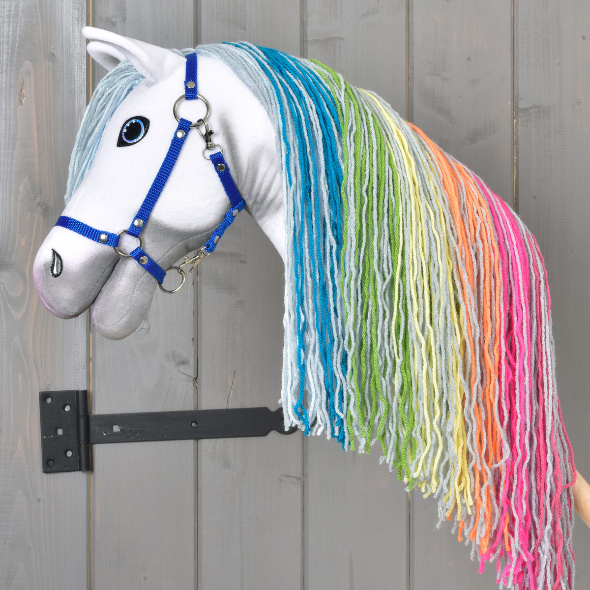 Hobby Horse Rainbow New od Hobby Horse LarDen s modrou uzdou a dlhou dúhovou priadzovou hrivou visí na nástennom držiaku na drevenom pozadí a dodáva miestnosti rozprávkové čaro