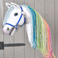 Hobby Horse Rainbow New od Hobby Horse LarDen má biele telo, dlhú dúhovú hrivu z priadze a modrú ohlávku pre rozprávkový vzhľad, na sivom drevenom obklade s čiernym kovovým závesom vľavo
