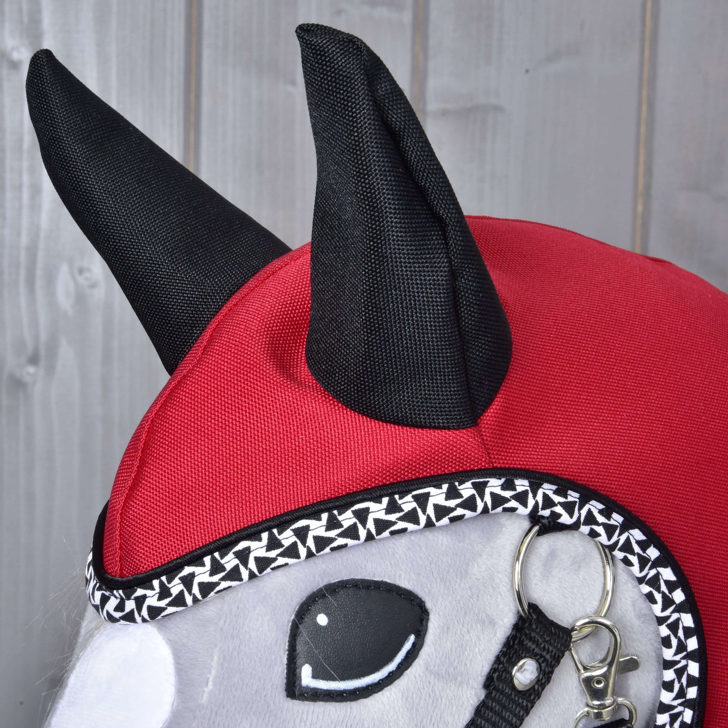 Detail plyšovej hračky Hobby Horse LarDen „Pláštenka Red Ferrari“ s červeno-čiernou kapucňou so špicatými ušami, ozdobným lemom, uzdečkou a vyšívanými očami, pripomínajúcou pláštenku do každého počasia na drevenom pozadí.