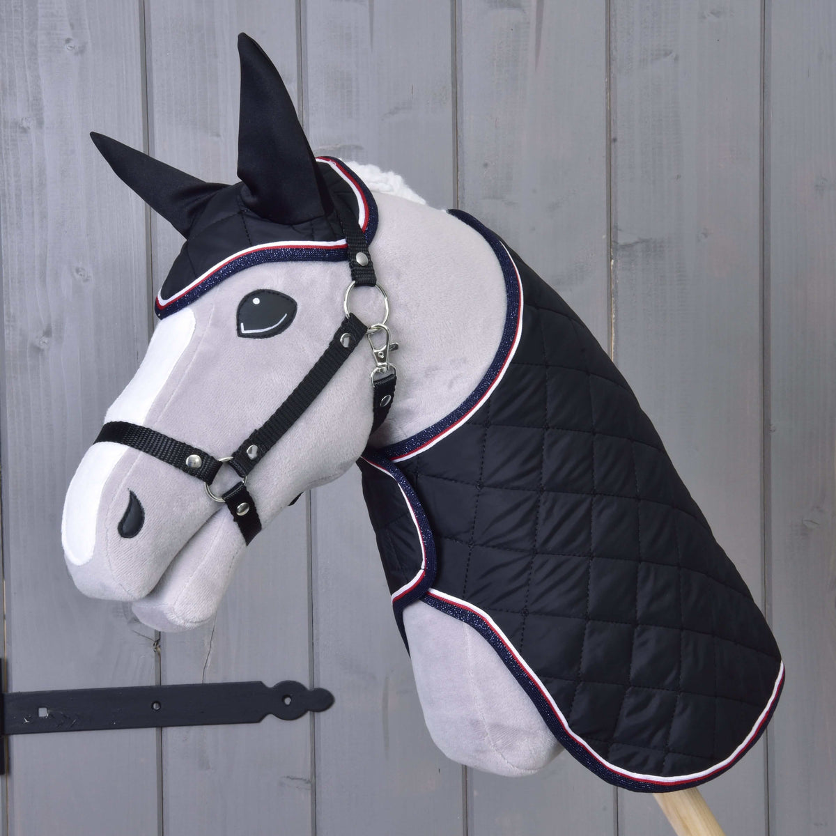 Sivá plyšová figurína koňa predvádza Set Royal od Hobby Horse LarDen, čiernu prešívanú deku a ladiacu čabraku. V pozadí je svetlá drevená stena s čiernou kovovou zástrčkou vľavo.