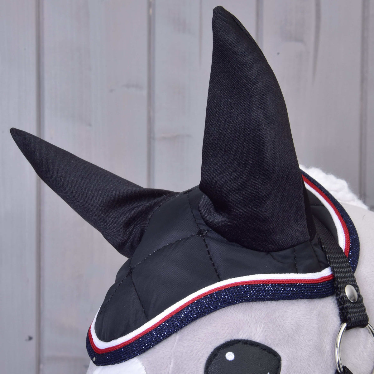 Detail Set Royal od Hobby Horse LarDen: hlava Hobby Horse s čiernymi látkovými ušami, štýlová čabraka s červeným, bielym a modrým lemom, sivá tvár a čierna uzdečka na pozadí svetlého dreva.