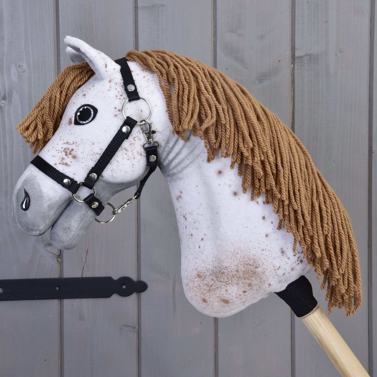 Detail Hobby Horse Rufus s čiernou ohlávkou od Hobby Horse LarDen s bielou tvárou, hnedou hrivou z priadze, čiernou ohlávkou a hnedými škvrnami, na pozadí drevených panelov.