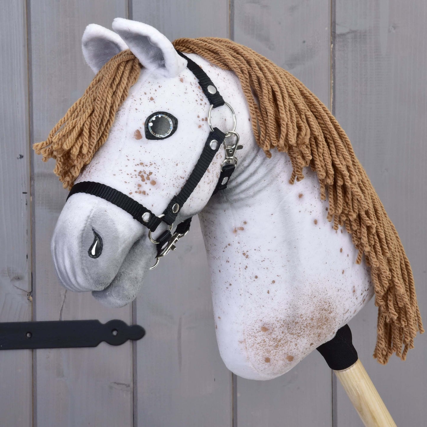 Hobby Horse Rufus s čiernou ohlávkou od Hobby Horse LarDen s ručne vyrobenou bielo škvrnitou látkovou hlavou, hnedou priadzou ako hrivou, gombíkovými očami a čiernou ohlávkou na drevenej tyči, zobrazený na pozadí sivého dreva.