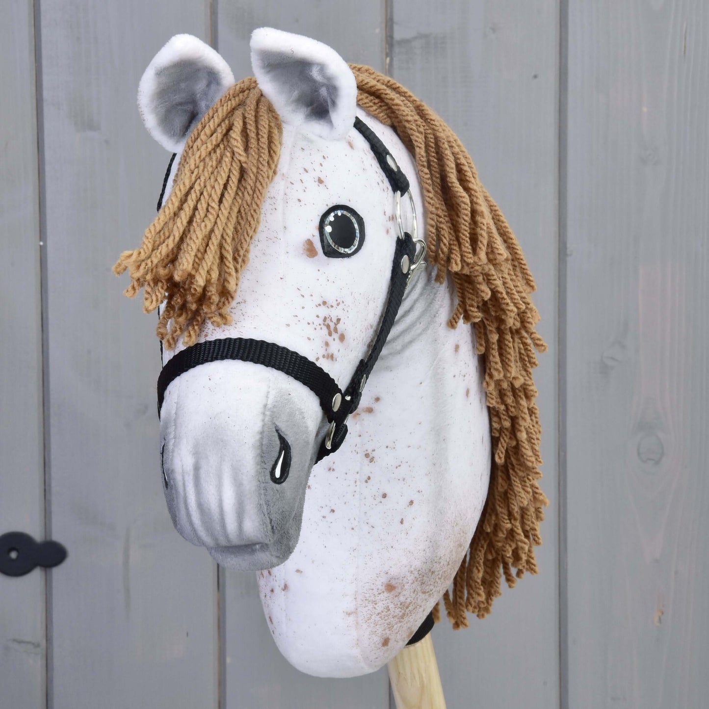 Hobby Horse Rufus s čiernou ohlávkou od Hobby Horse LarDen s ručne vyrobenou bielou plyšovou tvárou, hnedou hrivou z priadze, čiernou ohlávkou, maľovanými detailmi a upevnený na drevenej tyči na pozadí sivých drevených panelov.