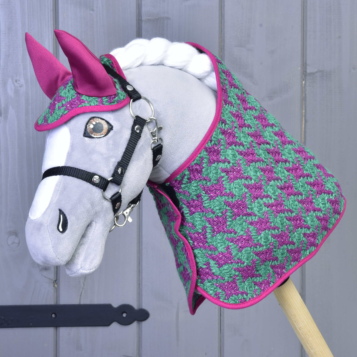 Sivý Hobby Horse LarDen pred svetlo sivou drevenou stenou, na sebe Set Coco: set deky v ružovo-zelenom vzore kohútej stopy s ladiacou ružovou čabrakou na uši a čiernou uzdečkou.