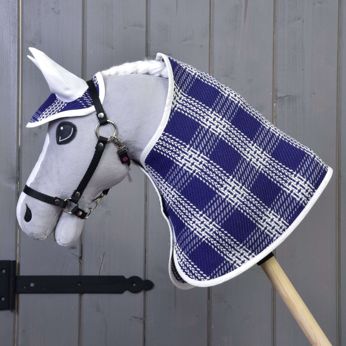Hobby Horse LarDen „Set Blue Cube“ so sivou plyšovou hlavou, modro-bielou károvanou sadou deky s ladiacou čabrakou, upevnený na drevenej tyči pred sivou drevenou stenou.