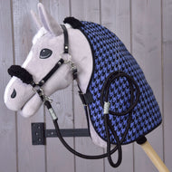 Plyšový Hobby Horse Set Harmony Blue od Hobby Horse LarDen s čiernou ohlávkou, opratami a modro-čiernou stajňovou dekou so vzorom kohútej stopy je pripevnený na stene na pozadí svetlých drevených dosiek.