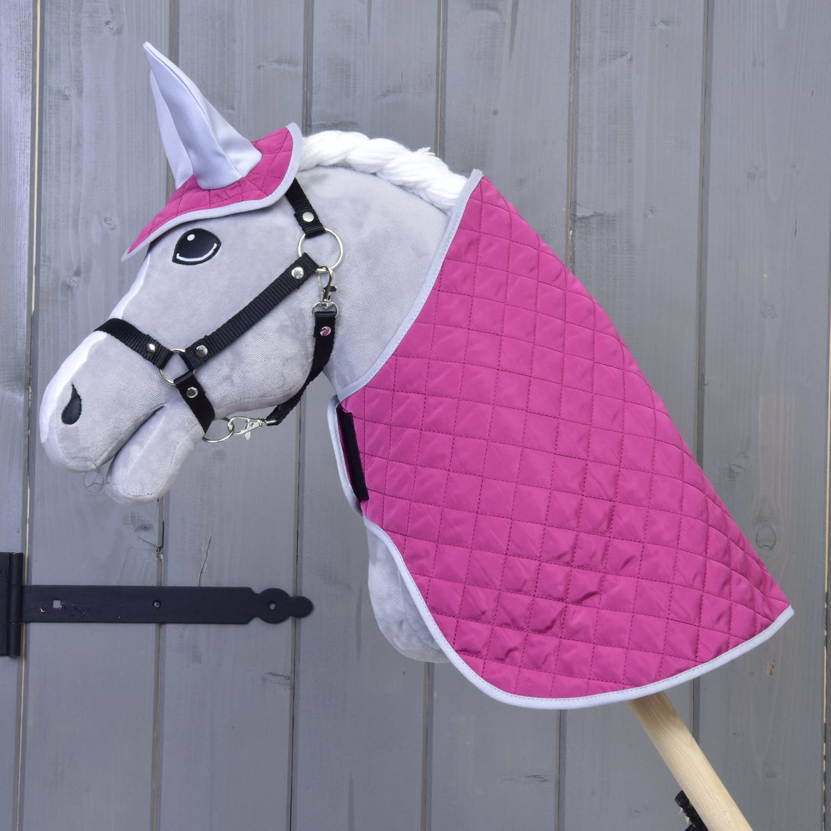 Set Hot Pink Silver od Hobby Horse LarDen: plyšový Hobby Horse so sivou tvárou, čiernou uzdečkou, bielou hrivou, prešívaným setom deky v sýto ružovej a bielej farbe a ladiacou čabrakou na uši, všetko na drevenej tyči pred drevenou stenou.