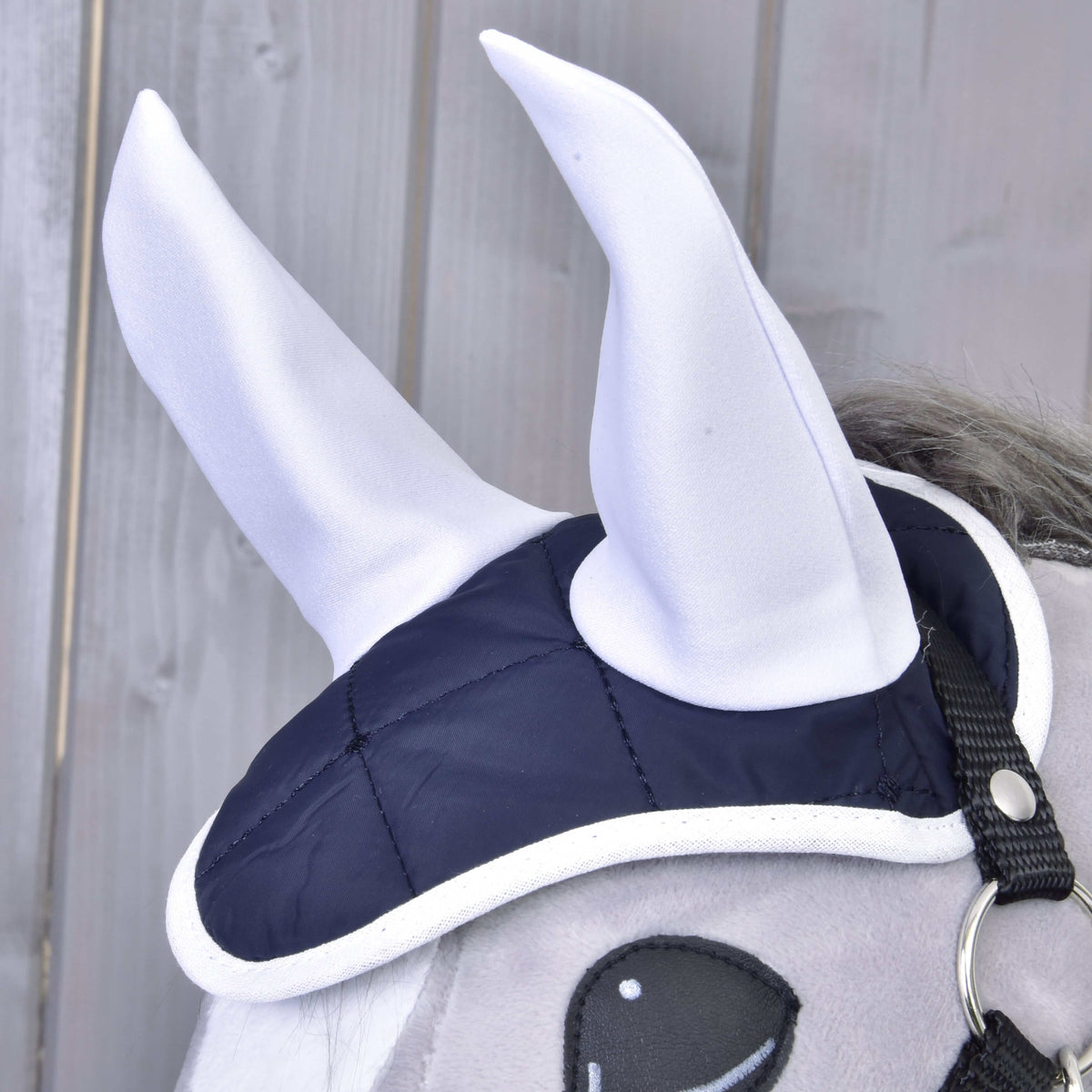 Set Monte od Hobby Horse LarDen: sivá plyšová konská hlava s tmavomodro-bielou sadou čabraky na uši s dvoma veľkými bielymi špicatými látkovými rohmi, zobrazená pred dreveným obložením pre výrazný vzhľad.