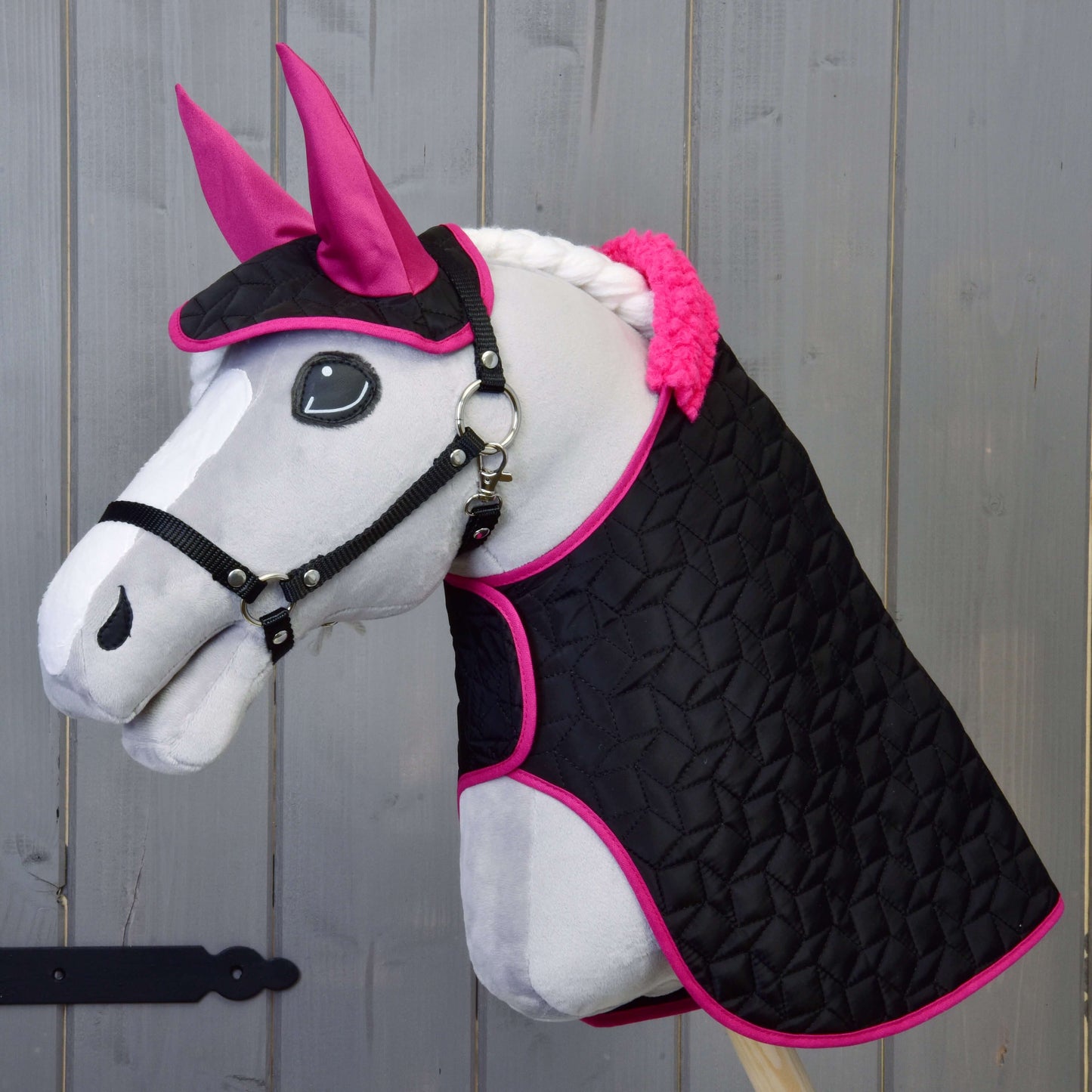 Hobby Horse LarDen "Set Thermo čierny s kožúškom" s plyšovým Hobby Horse s bielou tvárou, ružovými ušami, čiernou uzdečkou a spletenou bielou hrivou, na drevenom pozadí. Má čiernu prešívanú deku lemovanú jasne ružovou farbou.
