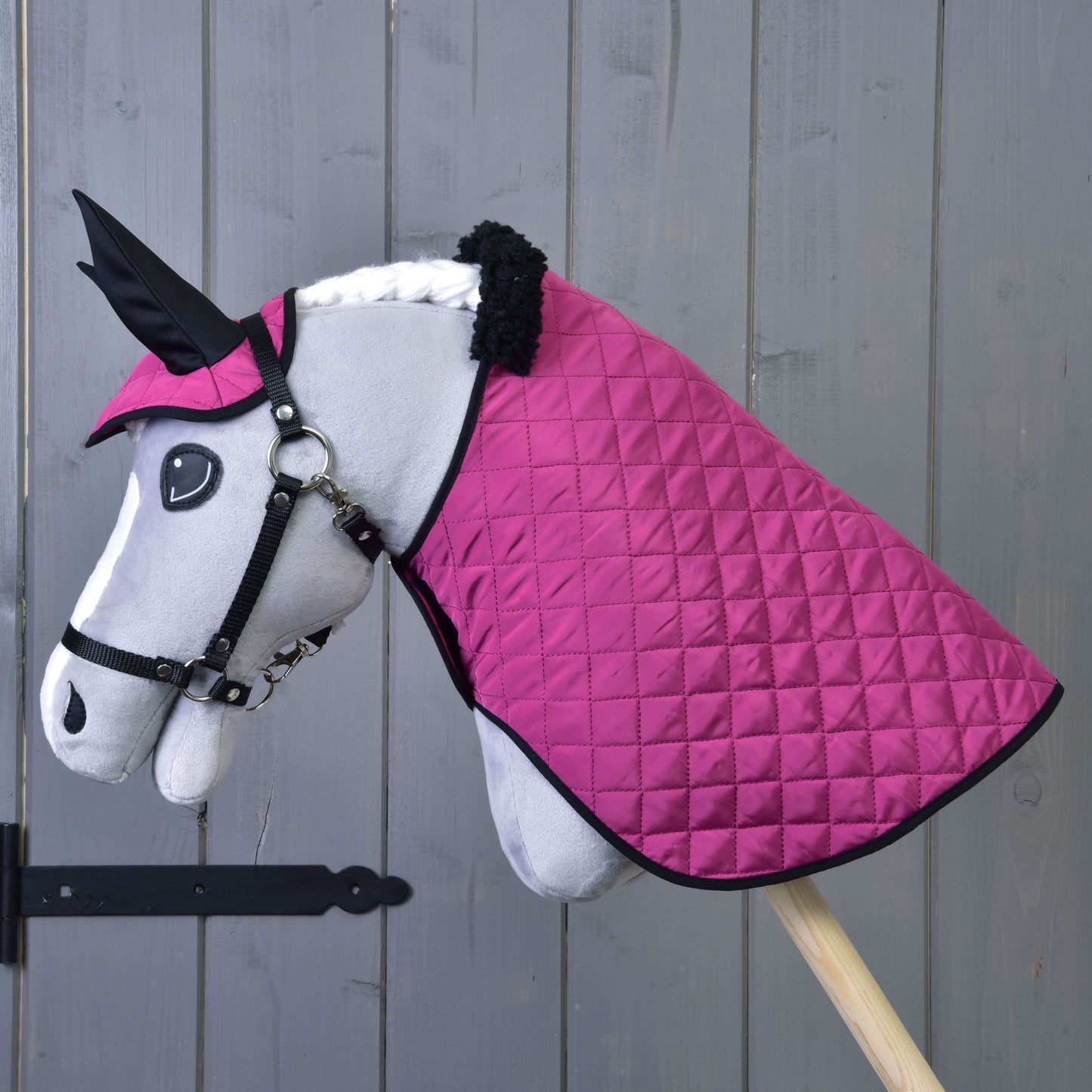 Plyšový Hobby Horse od Hobby Horse LarDen stojaci na drevenej tyči s bielou tvárou, čiernou uzdečkou a dekou a čabrakou Set Thermo Pink pred sivou stenou, ako pôvabná jazdecká dekorácia.