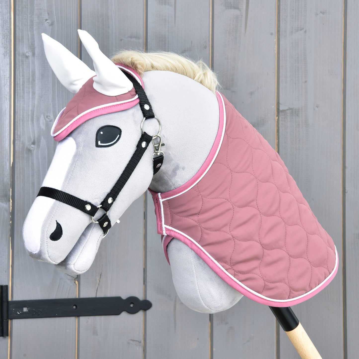 Hobby Horse LarDen „Set Dusty Rose“ na sivom Hobby Horse s bielou hrivou, čiernou uzdečkou, čabrakou, prešívanou ružovou dekou a čiernou tyčou, zobrazenom pred drevenou stenou.