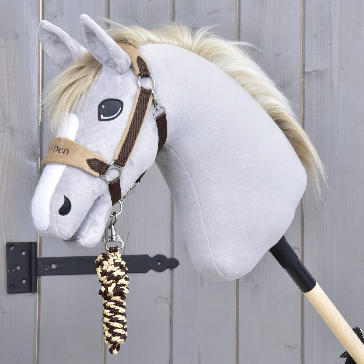 Set LarDen Exclusive Braun od Hobby Horse LarDen, sivý Hobby Horse s uzdečkou, fleecovým nánosníkom a krémovo hnedým pleteným vodidlom na drevenej tyči, odfotografovaný pred svetlosivou drevenou stenou.