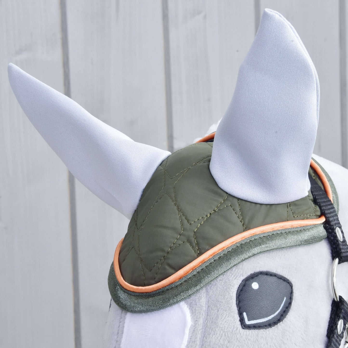 Detail plyšového Hobby Horse LarDen s čabrakou Sport, zelenou prešívanou čabrakou s oranžovým lemovaním a mäkkými vzpriamenými svetlosivými ušami, na svetlom drevenom pozadí.