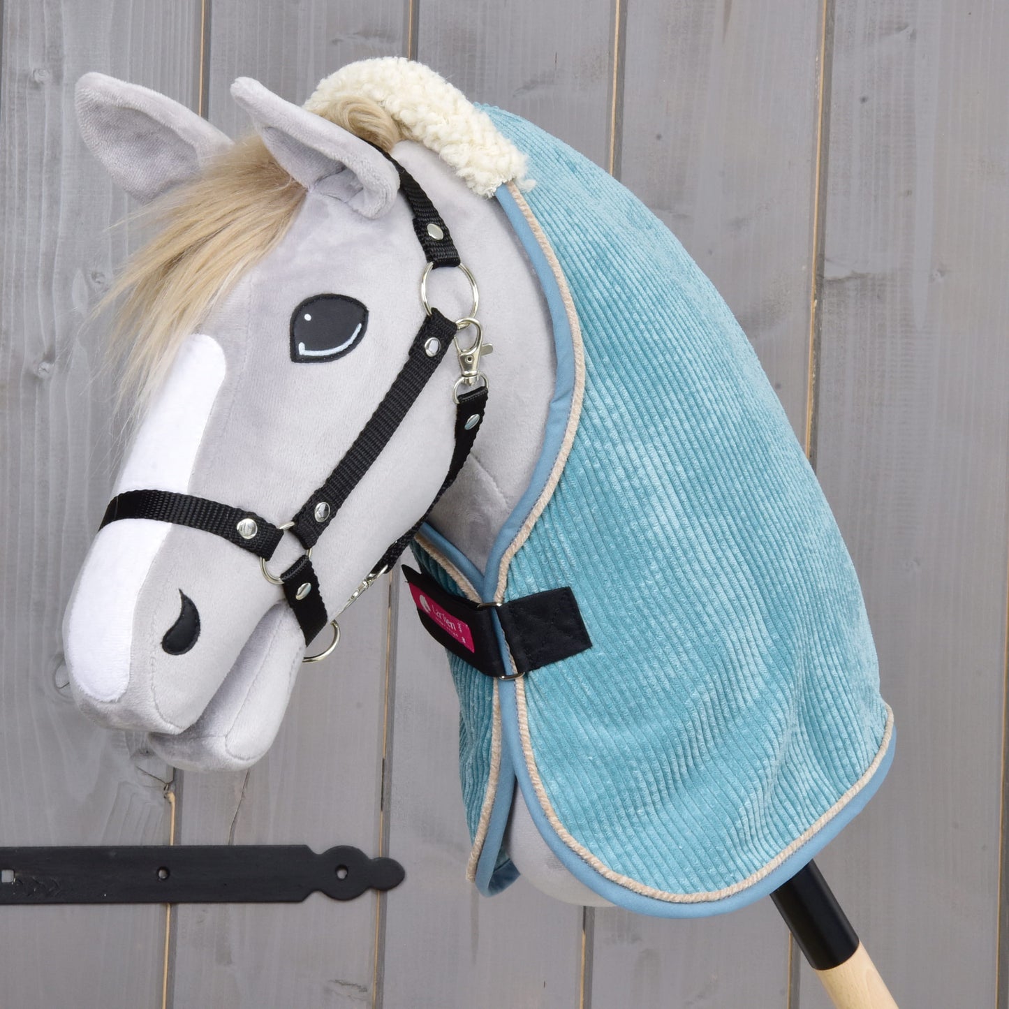 Plyšový Hobby Horse od Hobby Horse LarDen so svetlosivou tvárou, čiernou uzdečkou, bielou papuľou a dekou Deka stajňová Fern zo svetlomodrého materiálu PES, pózujúci na drevenom pozadí.
