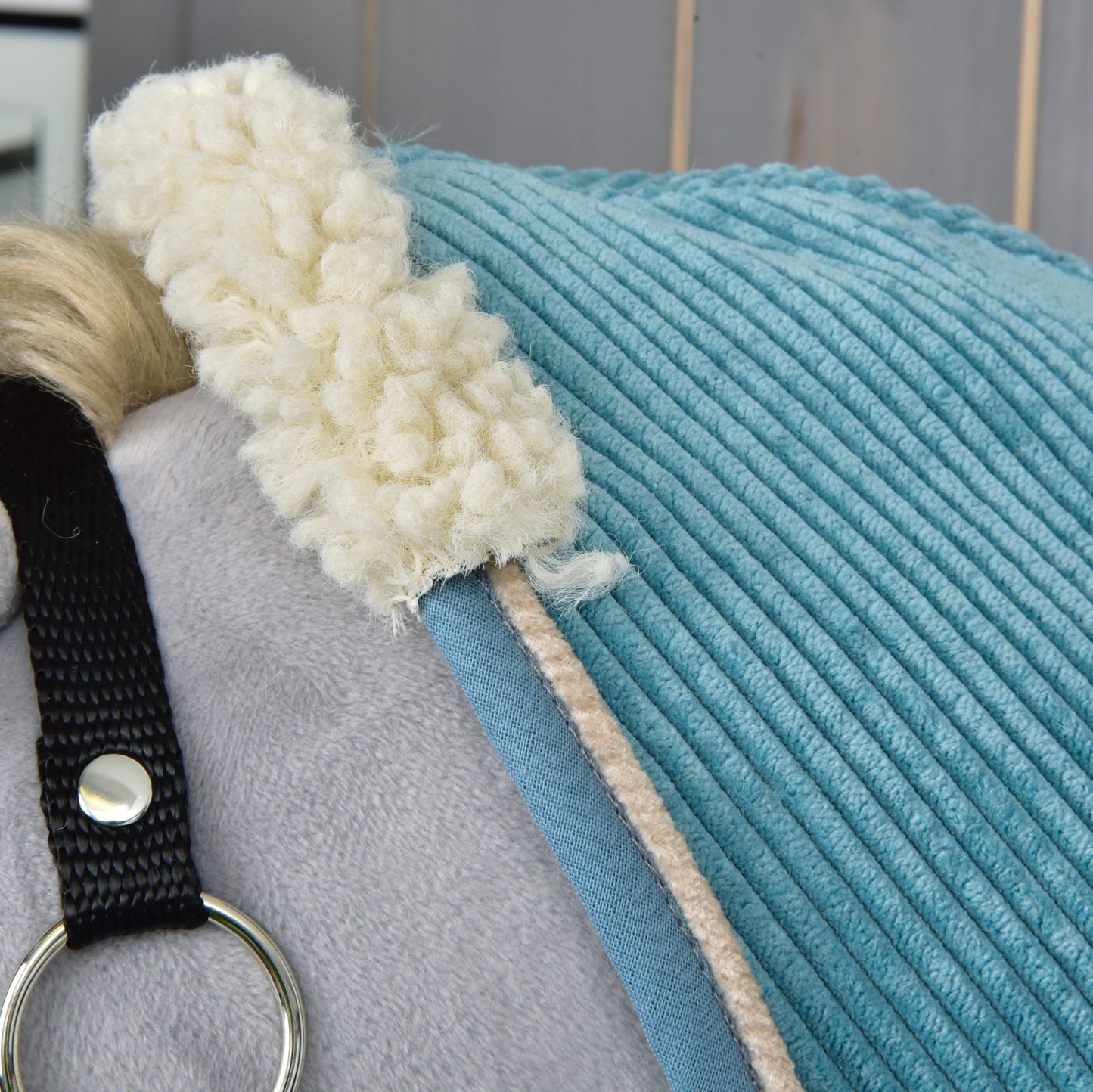 Detail svetlosivého plyšového koňa s modrou dekou Deka stajňová Fern od Hobby Horse LarDen, s krémovým flísovým lemovaním a čiernym pásikom so strieborným krúžkom. V pozadí vidno svetlé drevené panely.