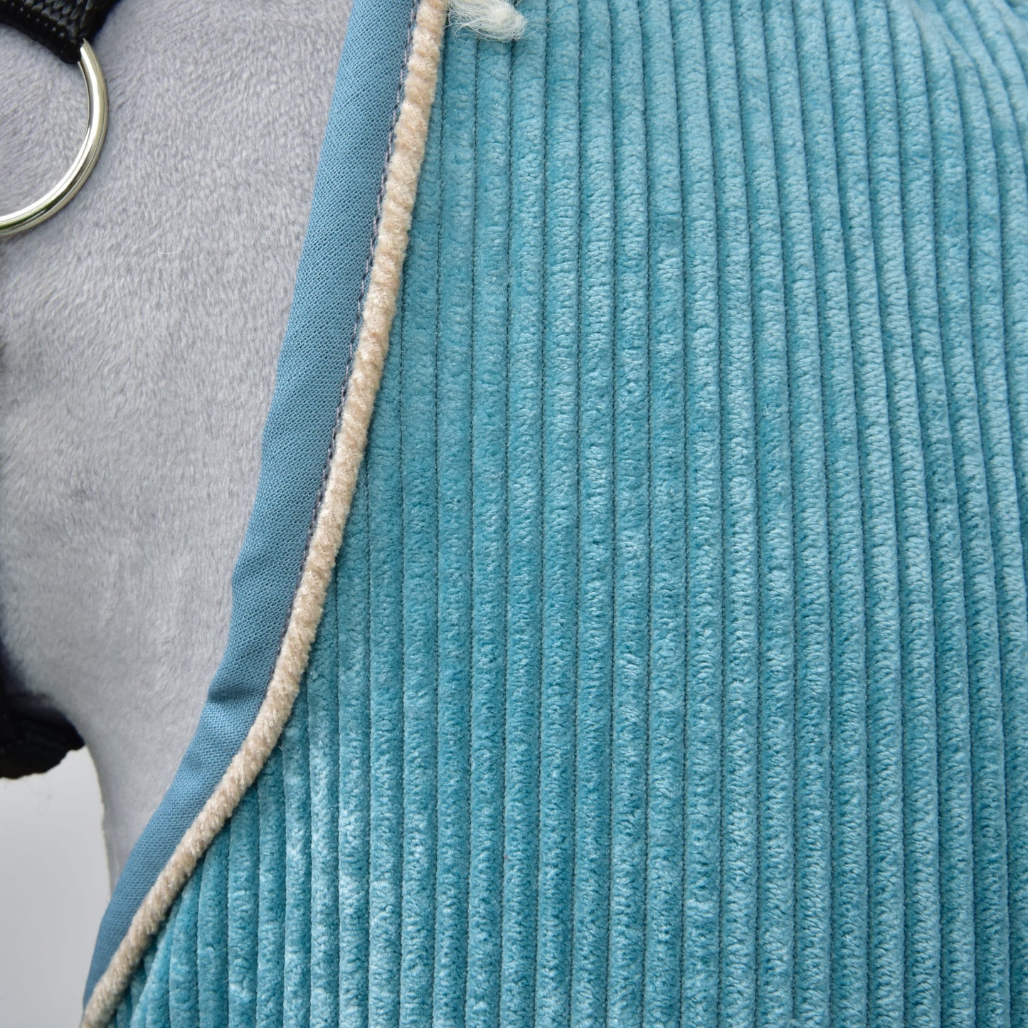 Detail textúrovanej modrej deky Deka stajňová Fern od Hobby Horse LarDen prehodenej cez sivého koňa, s béžovým chlpatým lemom a viditeľným kovovým krúžkom z konského postroja.