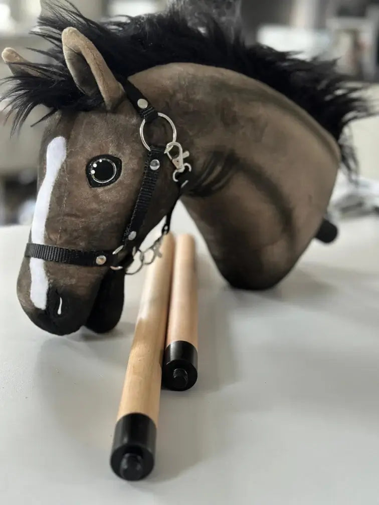 Plyšová hlava Hobby Horse s čiernou hrivou a ohlávkou leží na stole vedľa dvoch drevených tyčí s čiernymi koncovkami, ideálna pre nadšencov do Hobby Horsingu.