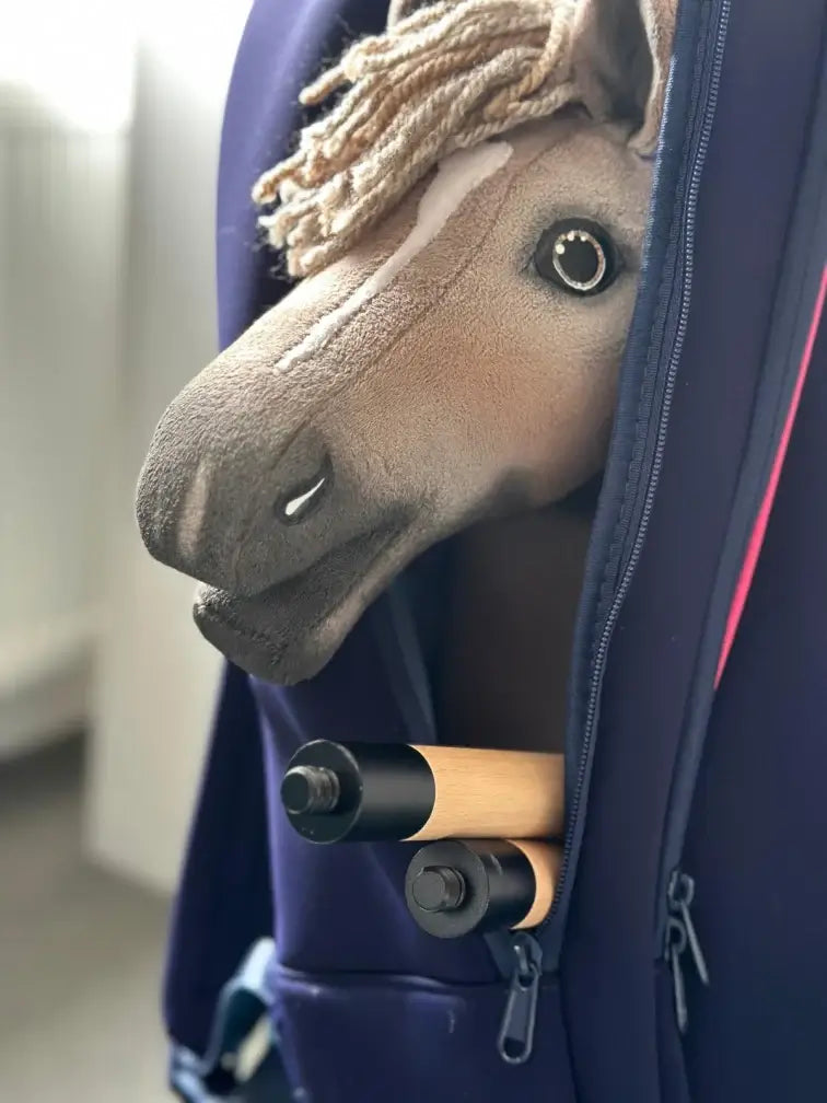 Plyšový Hobby Horse so svetlou hrivou a bielou lysinou vykúka z čiastočne rozopnutej tmavomodrej tašky, vnútri tašky vidno aj dve drevené tyče s držadlami — ideálny pre dobrodružstvá pri Hobby Horsingu.