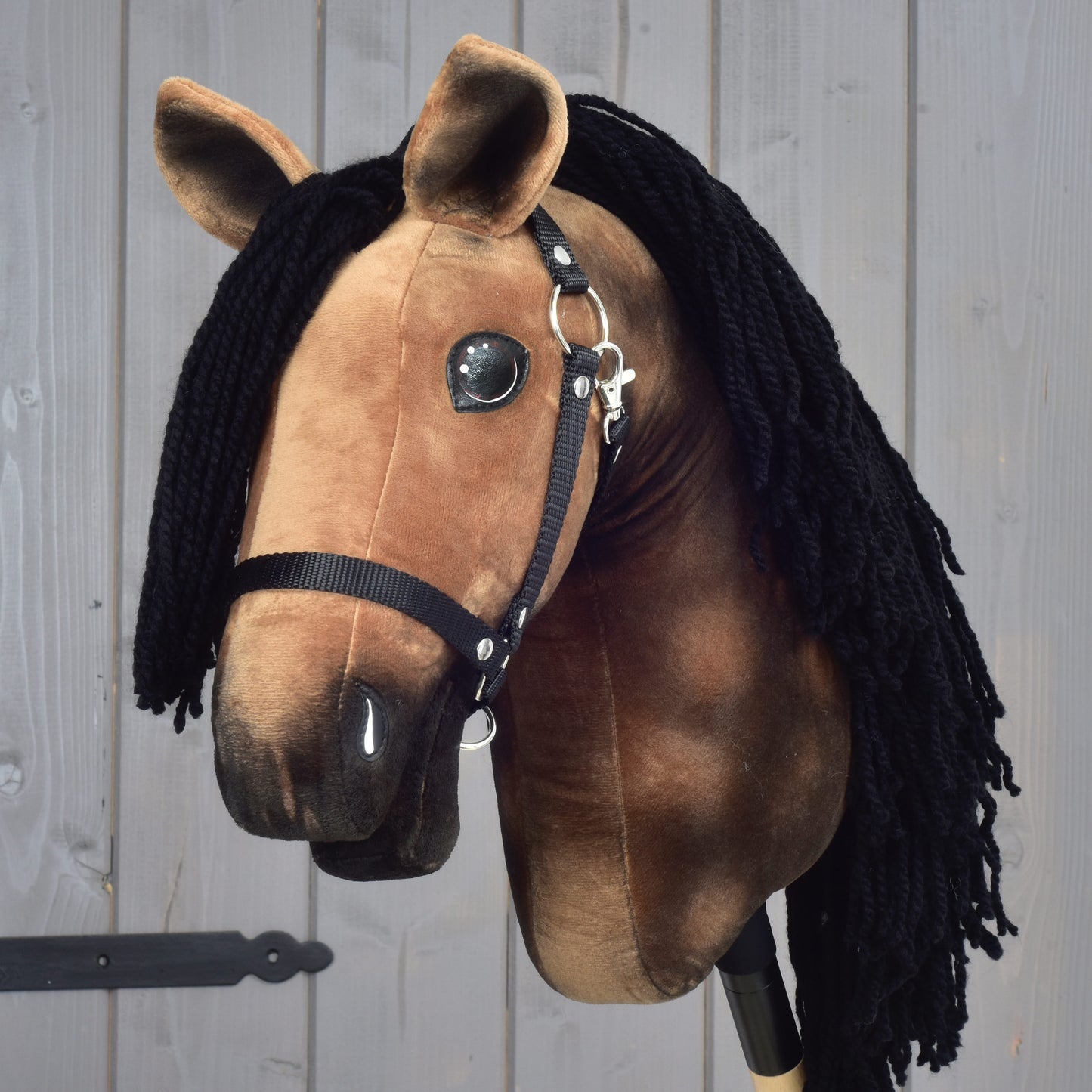 Hobby Horse Stormy s čiernou ohlávkou od Hobby Horse LarDen, s plyšovým hnedým telom a dlhou čiernou priadzovou hrivou, pred svetlosivou drevenou stenou.