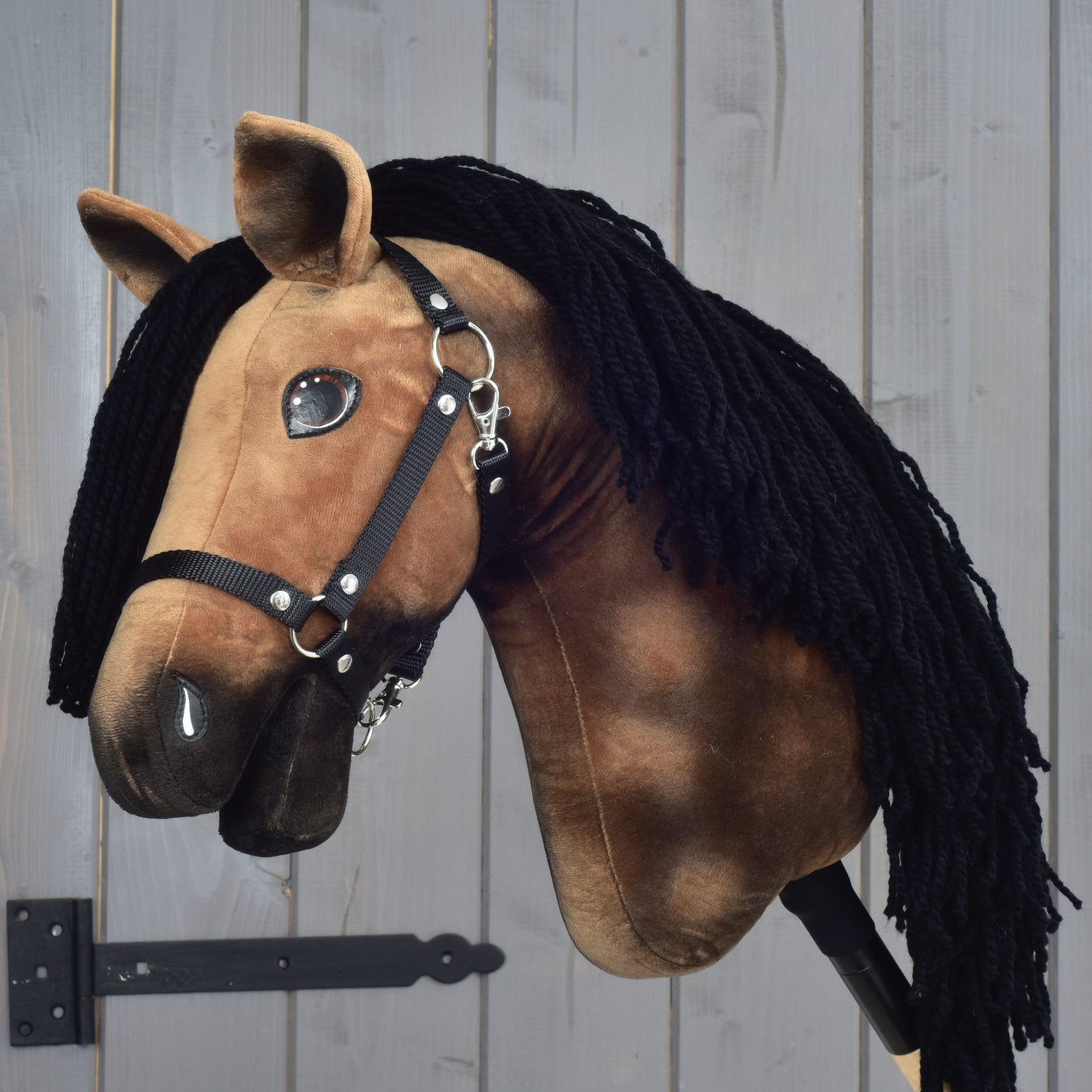 Hobby Horse Stormy s čiernou ohlávkou od Hobby Horse LarDen, hnedý plyšový model hannoverského koňa s čiernou priadzovou hrivou, čiernou ohlávkou a bielymi znakmi, nasadený na tyči na pozadí sivého dreva.