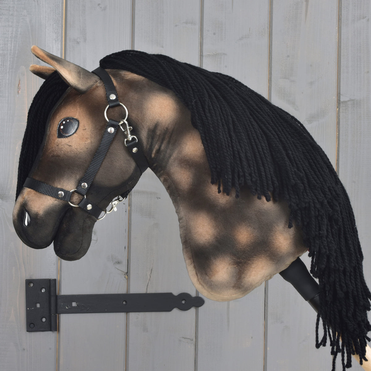 Realistický Hobby Horse Stormy with Black Halter od Hobby Horse LarDen, inšpirovaný hannoverským žrebcom a s čiernou priadzovou hrivou, je upevnený na sivej drevenej stene čiernym držiakom