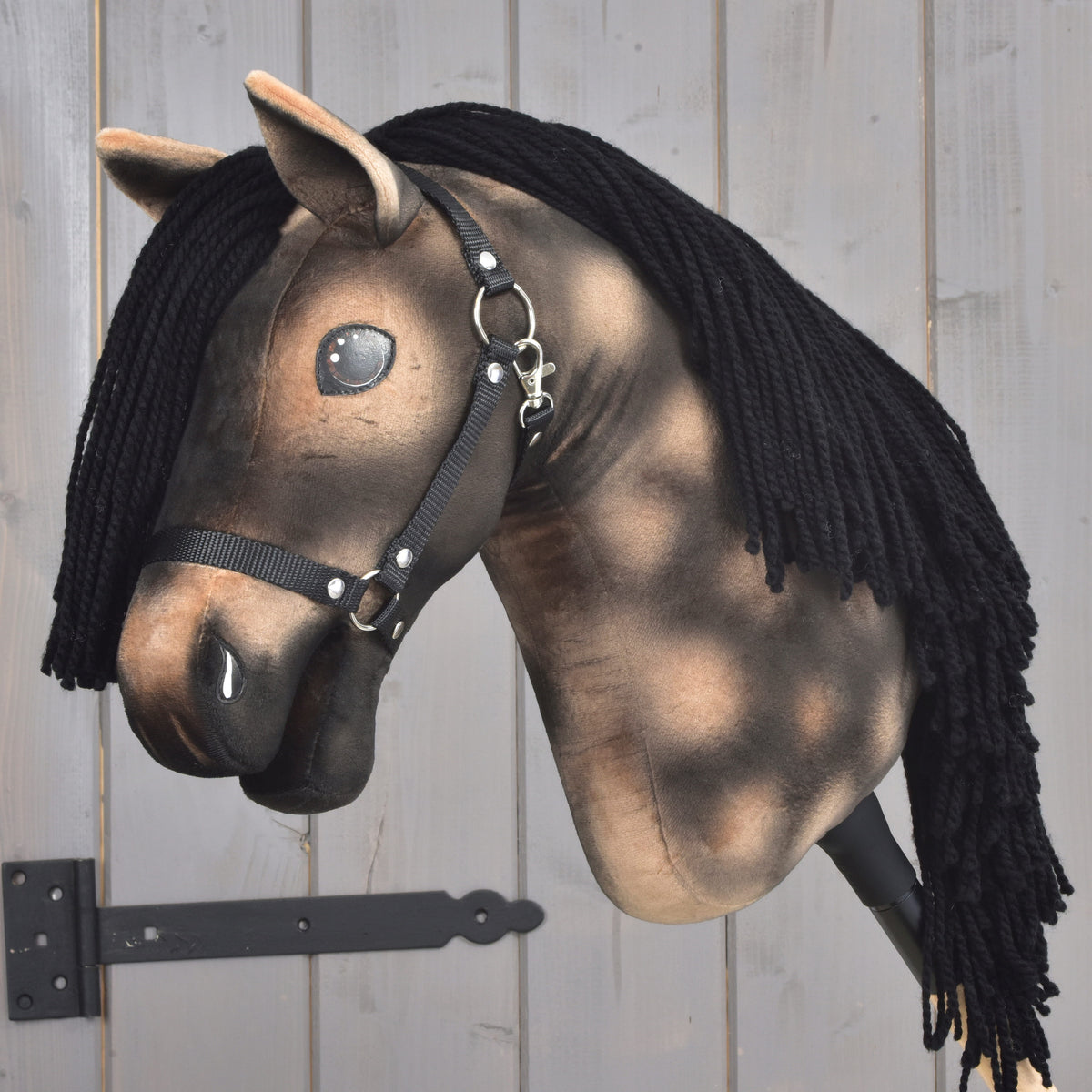 Hobby Horse Stormy od Hobby Horse LarDen má hnedú plyšovú hlavu, čiernu priadzovú hrivu, čiernu ohlávku a lesklé gombíkové oči, je na drevenej tyči pred svetlým dreveným pozadím a je inšpirovaný duchom hannoverského žrebca