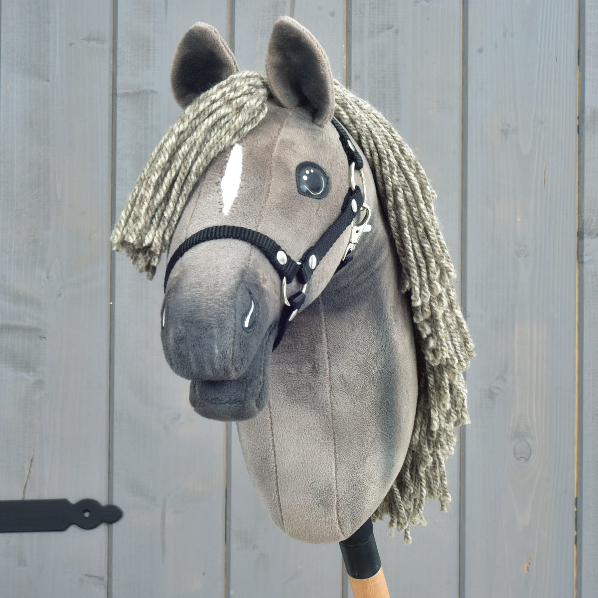 Hobby Horse Swing s čiernou ohlávkou - veľkosť S od Hobby Horse LarDen, detail elegantného sivého prevedenia s priadzovou hrivou, gombíkovými očami, výraznou bielou lysinou na tvári a čiernou ohlávkou, pripomínajúci vytríbený vzhľad hannoverského žrebca, na drevenom pozadí.