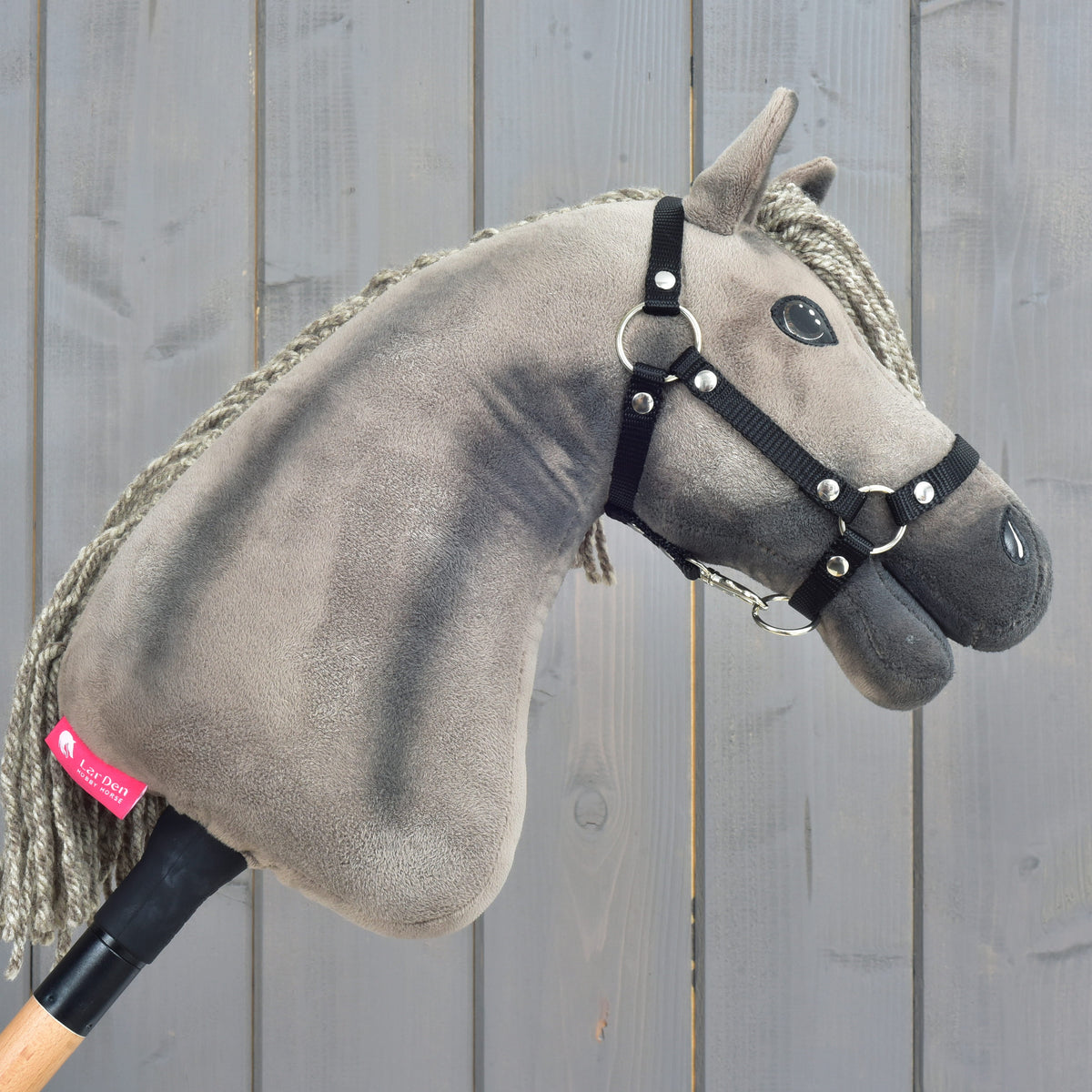 Hobby Horse Swing s čiernou ohlávkou - veľkosť S od Hobby Horse LarDen, s plyšovým prevedením, sivou hrivou a na drevenej tyči, zachytávajúci vznešený pôvab hannoverského žrebca, vystavený pred ošarpaným sivým dreveným pozadím.