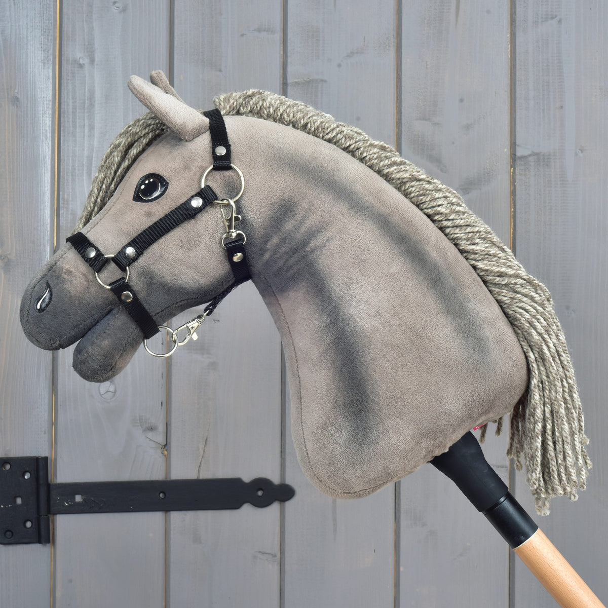 Hobby Horse Swing s čiernou ohlávkou - veľkosť S od Hobby Horse LarDen, sivý plyšový Hobby Horse pripomínajúci hannoverského žrebca, s čiernou ohlávkou a povrazovou hrivou, na drevenej tyči, vystavený pred drevenou stenou s viditeľnou kresbou dreva a čiernym kovovým závesom.
