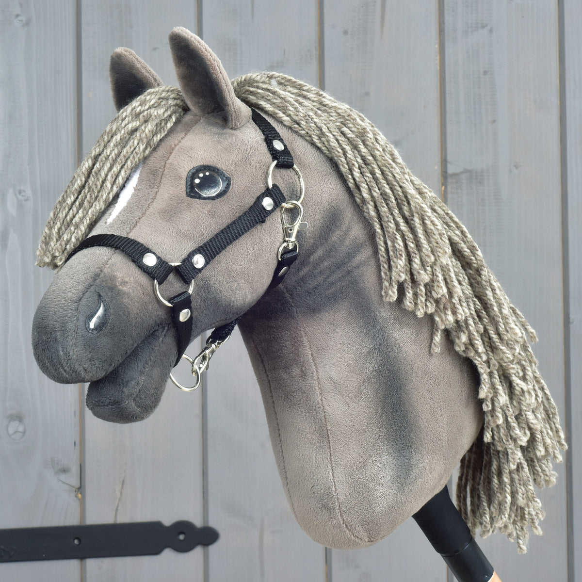 Hobby Horse LarDen "Hobby Horse Swing with black halter", veľkosť S, plyšový koník so zapletenou priadzovou hrivou a elegantnou čiernou ohlávkou, nasadený na tyči a vystavený pred dreveným pozadím.