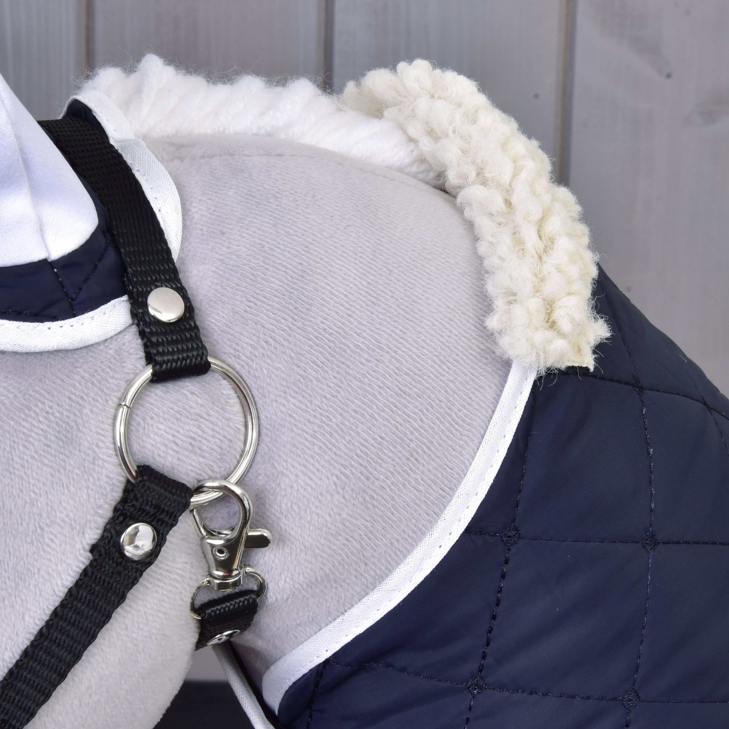 Detail psieho ramena v kabátiku Set Thermo Blue od Hobby Horse LarDen s prešívanou tmavomodrou látkou, bielym lemovaním a huňatým golierom, doplnený čiernym postrojom s kovovým krúžkom. V pozadí vidno drevené obloženie.
