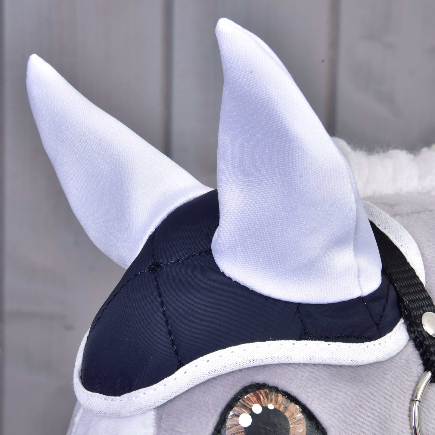 Detail hlavy plyšového koňa v elegantnom Set Thermo Blue od Hobby Horse LarDen, s bielymi ušami, tmavomodro-bielou čiapkou a čiernou uzdečkou. Perfektný dekoratívny kúsok na pozadí svetlých drevených panelov.