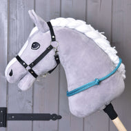 Nákrčník Tyrkys od Hobby Horse LarDen na sivom plyšovom Hobby Horse s bielou priadzovou hrivou, čiernou uzdečkou, modrými povrazovými opratami a ručne vyrobeným tyrkysovým cordeom na jazdenie bez uzdečky, na tyči pred drevenou stenou.