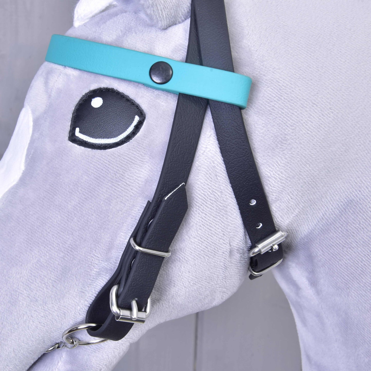 Detail uzdečky bez nánosníka tyrkysová s 2 čelenkami (veľ. M) od Hobby Horse LarDen na svetlosivom plyšovom koni s vyšívaným okom v kreslenom štýle. Uzdečka má strieborné pracky a vymeniteľnú čelenku, bez nánosníka, v tyrkysovej a čiernej farbe.