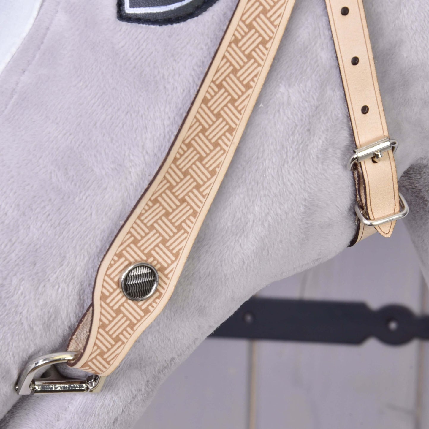 Detail Hobby Horse LarDen Uzdečky western Classic: svetlohnedý popruh z prémiovej kože s geometrickým vzorom a kovovým drukom, zapnutý na svetlosivom plyšovom povrchu, na rozmazanom pozadí.
