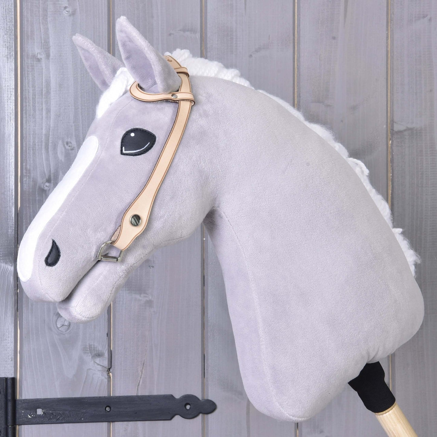 Uzdečka western jednouška od Hobby Horse LarDen na sivom plyšovom koni na drevenej tyči, s prémiovou koženou westernovou uzdečkou one-ear, na pozadí steny z drevených dosiek.
