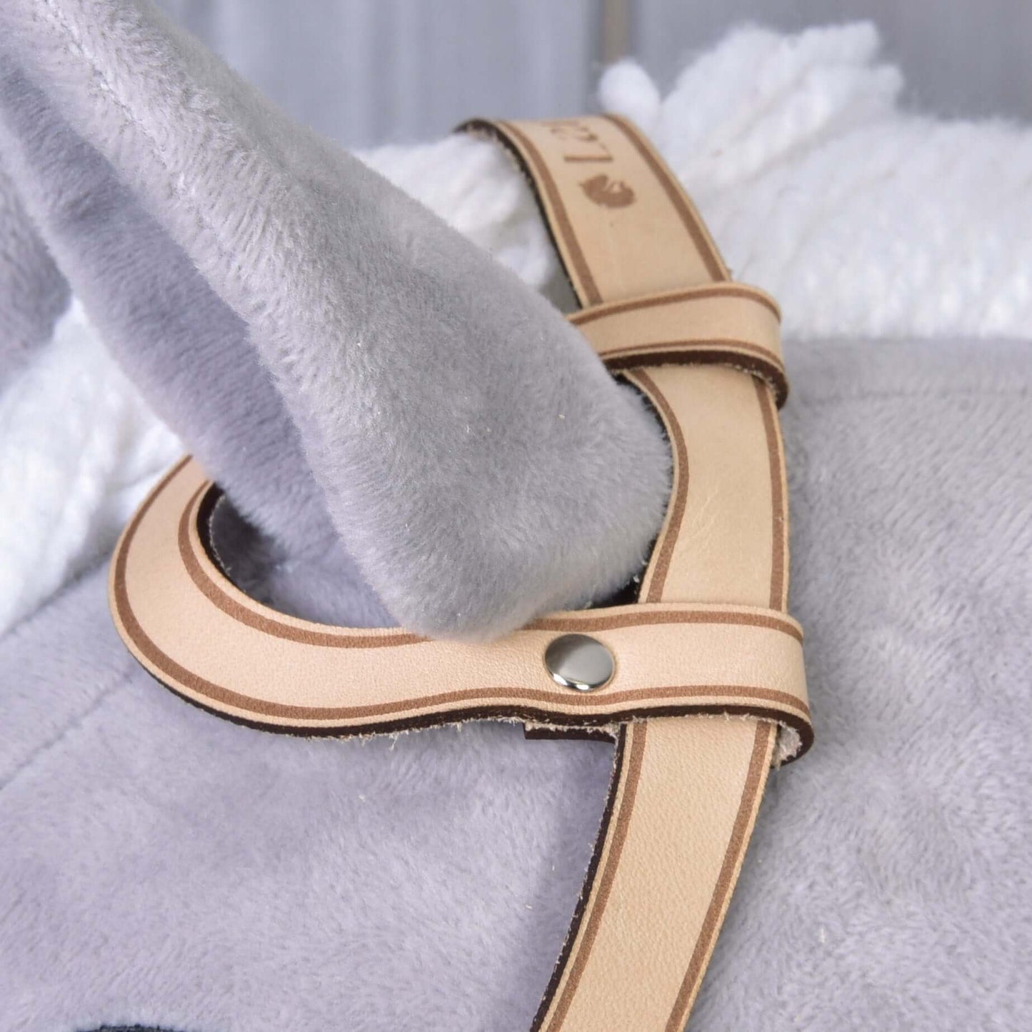 Detail sivej plyšovej látky a mäkkej bielej deky s uzdečkou western jednouška od Hobby Horse LarDen – svetlohnedým remienkom z kvalitnej kože s kovovými nitmi, prevlečeným okolo ovisnutého sivého plyšového ucha.