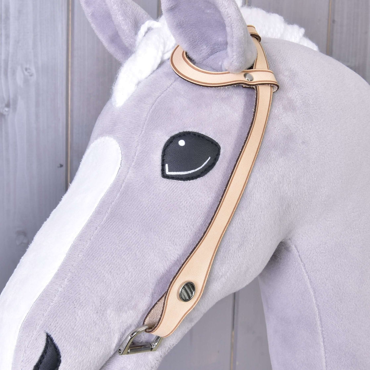 Detail hlavy plyšového koňa zo svetlosivej a bielej látky s vyšívaným čiernym okom a uzdečkou western jednouška od Hobby Horse LarDen so strieborným kovaním na drevenom pozadí.