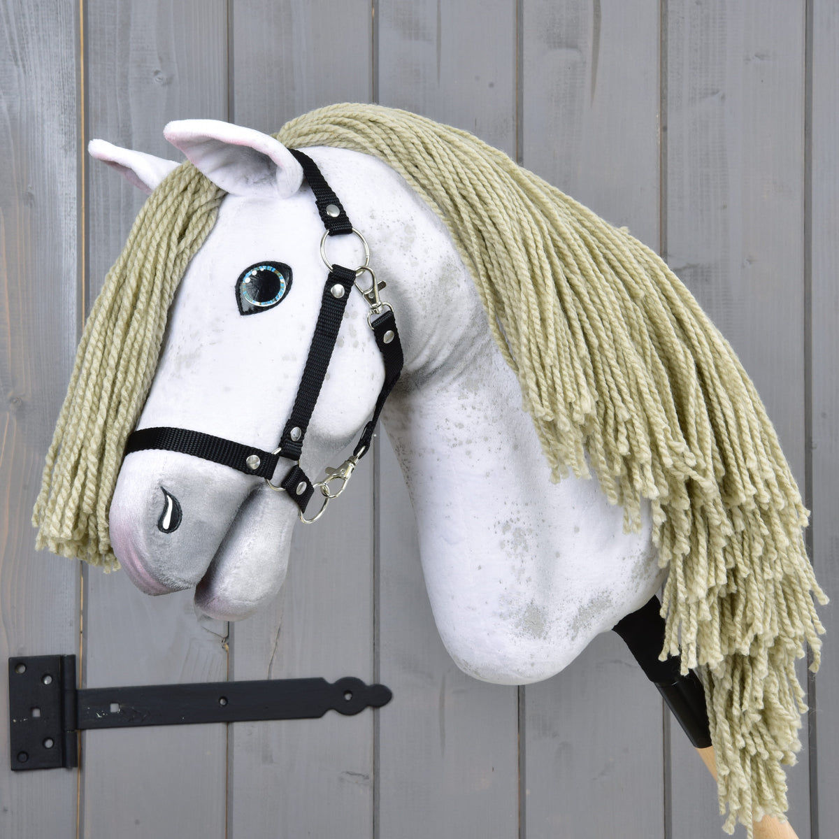 Hobby Horse Victory s čiernou ohlávkou - veľkosť M od Hobby Horse LarDen, s bielou plyšovou hlavou, blond hrivou z priadze, čiernou ohlávkou a maľovaným modrým okom, na drevenej tyči, pred sivou drevenou stenou.